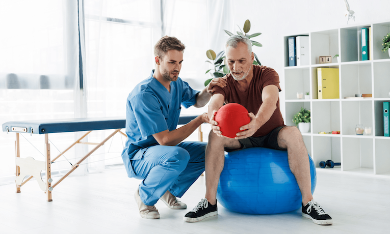 starší muž cvičí s fyzioterapeutom na fit lopte